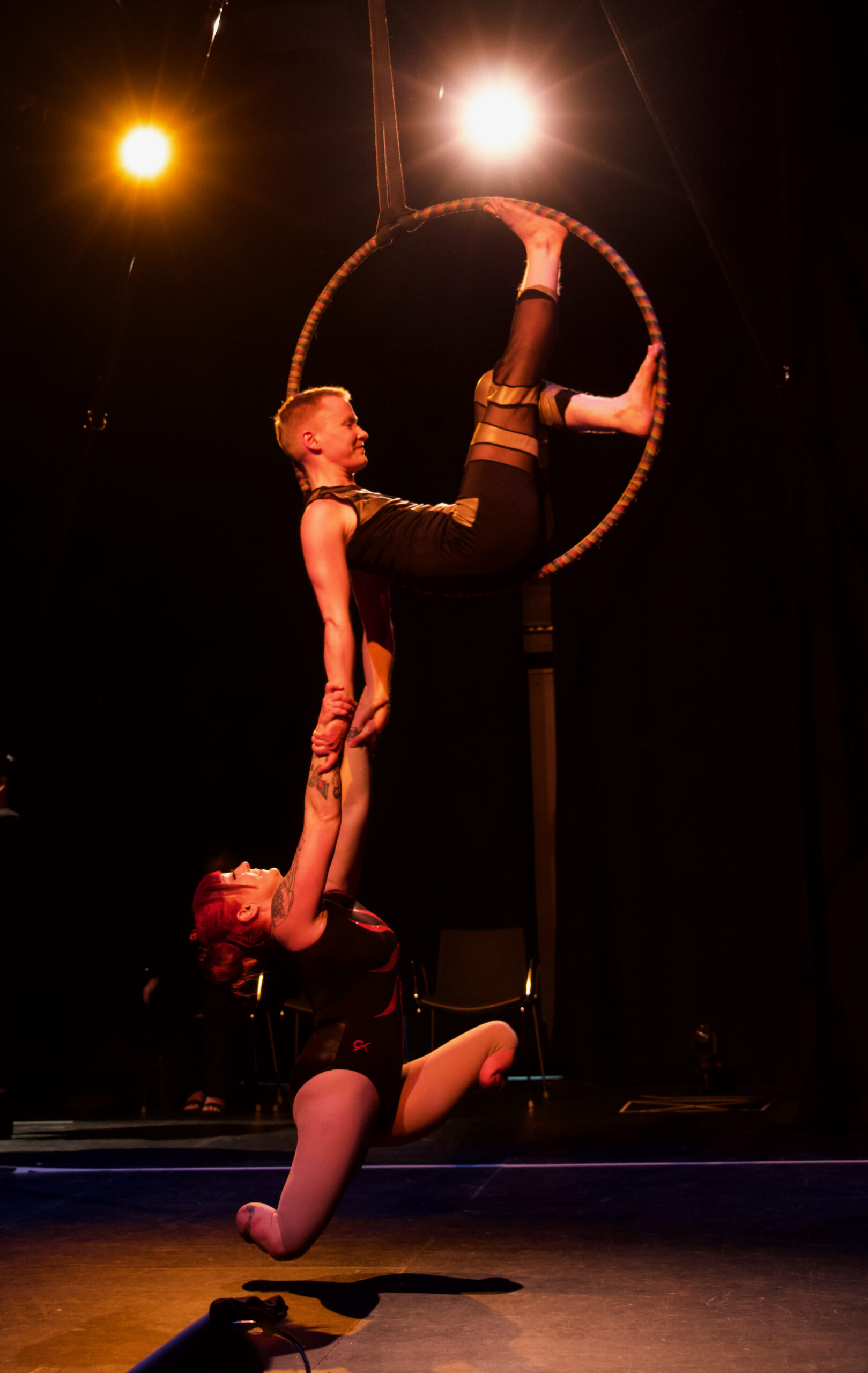 Onstage with low lighting, two white Disabled performers are in the air, on a suspended aerial hoop the size of an adult hula hoop at an average standing head height above the floor. Inside the hoop, Maxime sits side-profile, one leg up, leaning back into the curve of the hoop. Their arms hang straight down and holding onto them is Erin, a double below knee amputee. Erin is suspended, wearing no prosthetic legs, back arched like a crescent moon, and legs split apart. The two artists are smiling happily, celebrating their Queer Disabled Love. Jay Middaugh